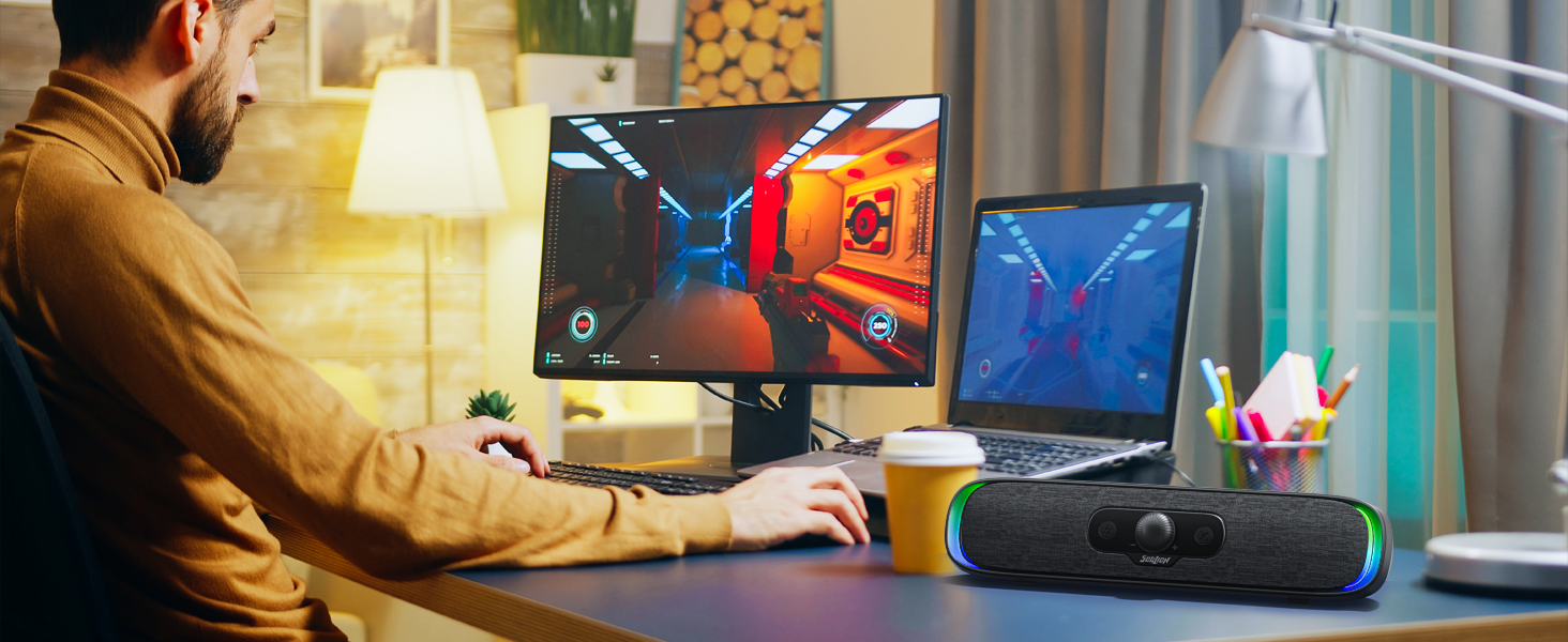 Gaming-Setup mit zwei Monitoren, auf denen farbenfrohe Spielszenen angezeigt werden. Der Desktop umfasst einen Laptop, einen kabellosen Lautsprecher und eine gelbe Tasse. Person in braunem Hemd, mit der Tastatur sichtbar.